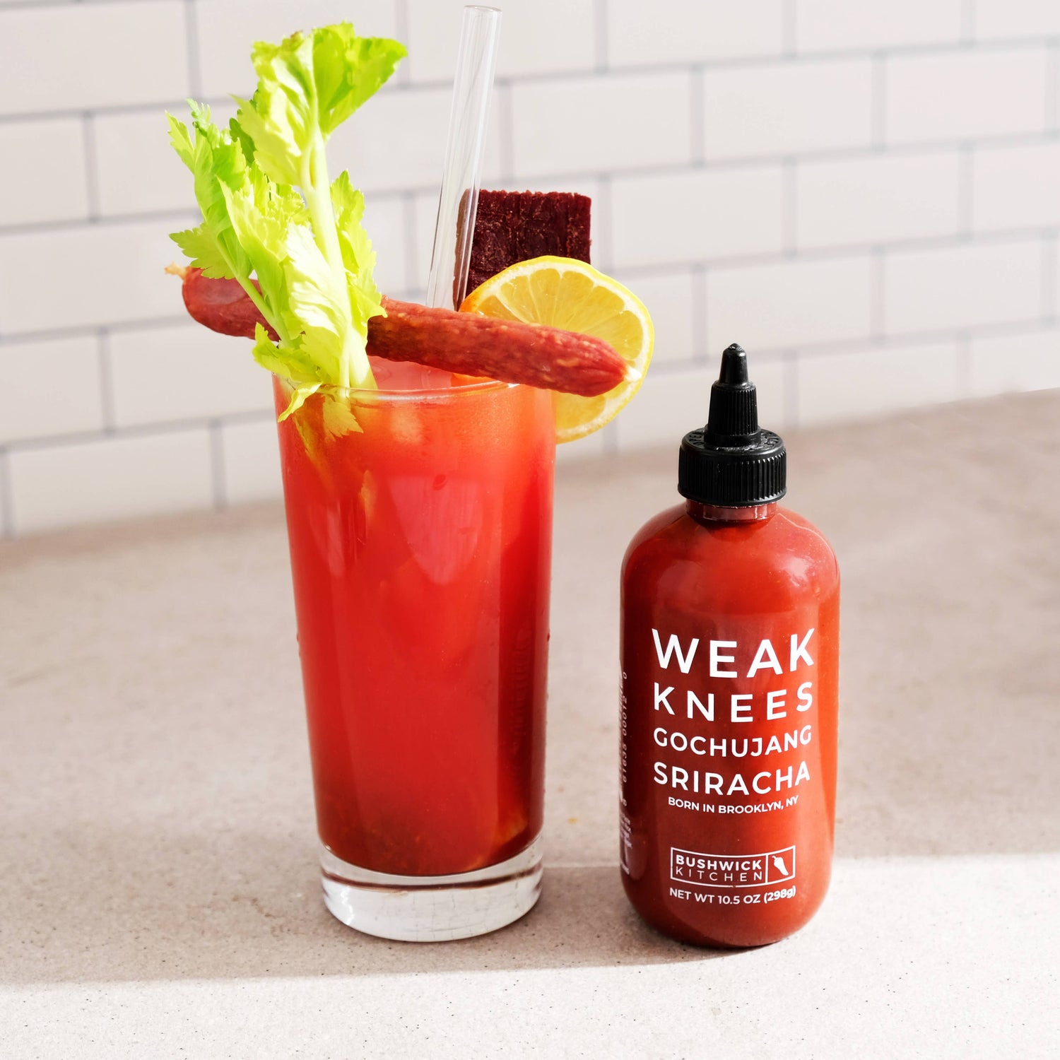 Bloody Mary cocktail with garnishes and a bottle of Weak Knees Cochujang Sriracha on a countertop.