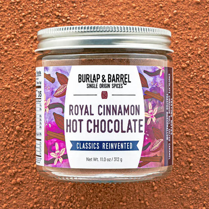 A jar of Burlap &amp; Barrel Royal Cinnamon Hot Chocolate on a textured brown background. The jar has a silver metal lid and a white label with purple and pink floral designs, set against a matching brown powder background.