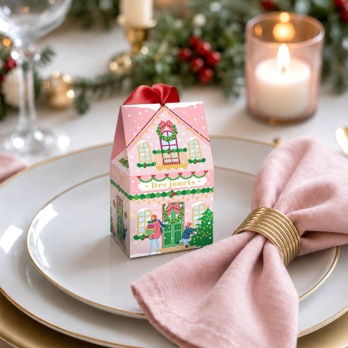 A small pink house-shaped gift box with a red ribbon sits on a white plate set on a dining table. The box is decorated with a colorful Christmas village illustration and the French text &