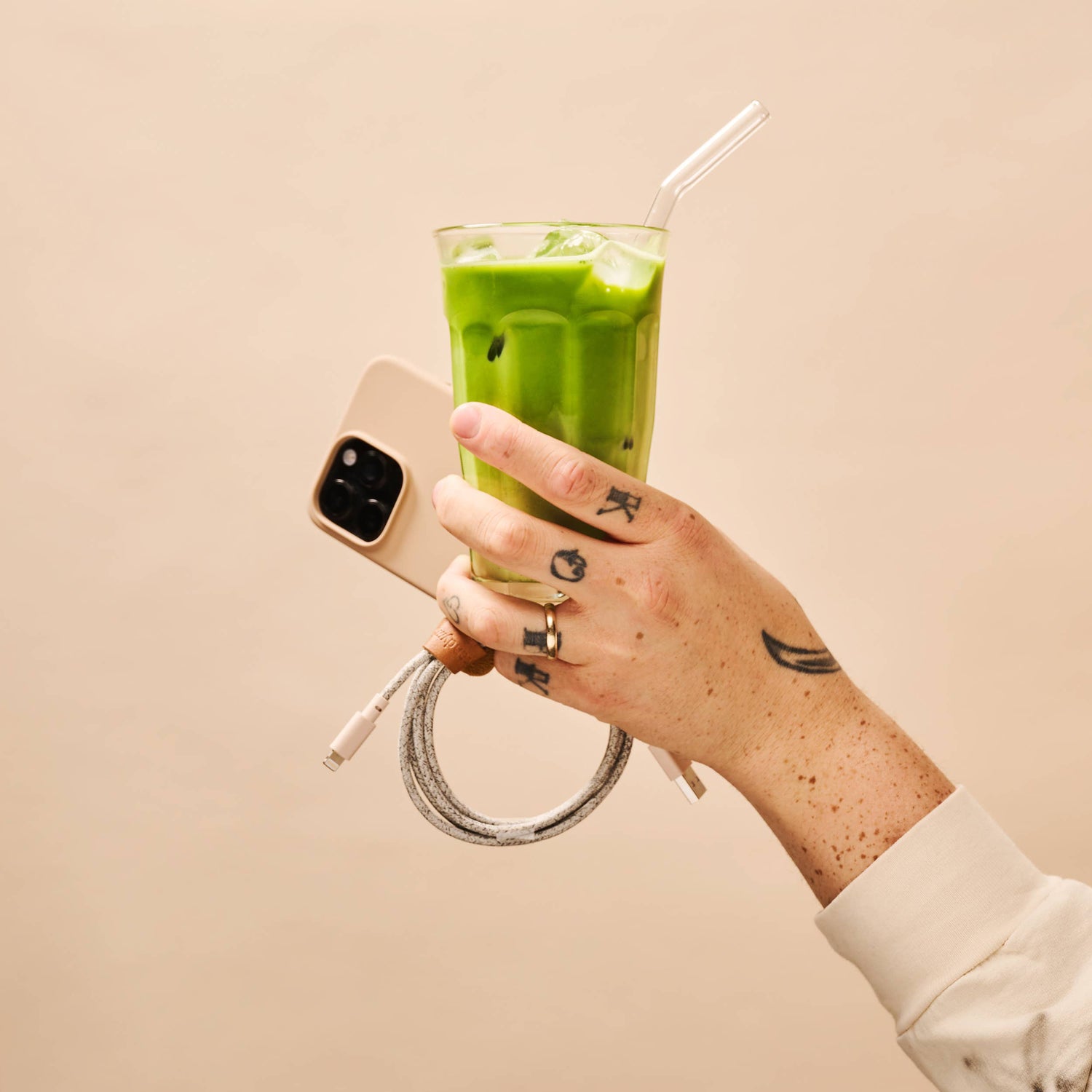 Hand holding a green smoothie with a straw, wearing a white sleeve against a beige background