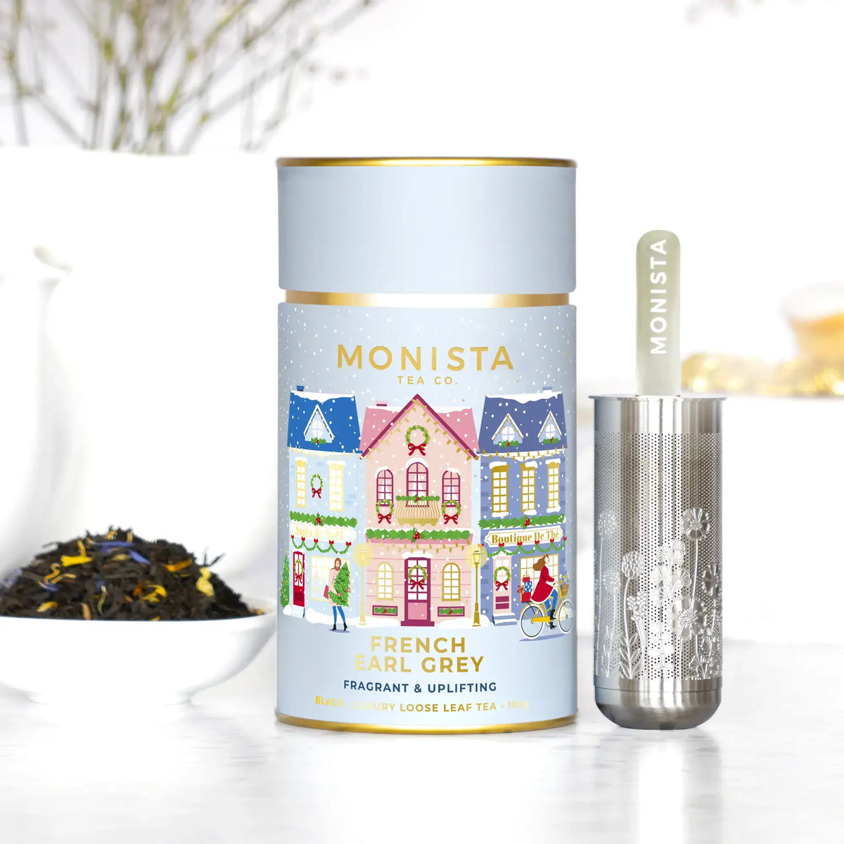 Monista Tea Co. French Earl Grey tea set featuring a pastel holiday/Christmas illustrated tin canister, a silver metal infuser, and a small bowl of loose leaf tea on a white surface.