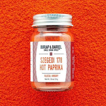 A glass jar of Burlap &amp; Barrel Szegedi 178 Hot Paprika spices, featuring a white label with red text, set against a vibrant textured background of the spice.