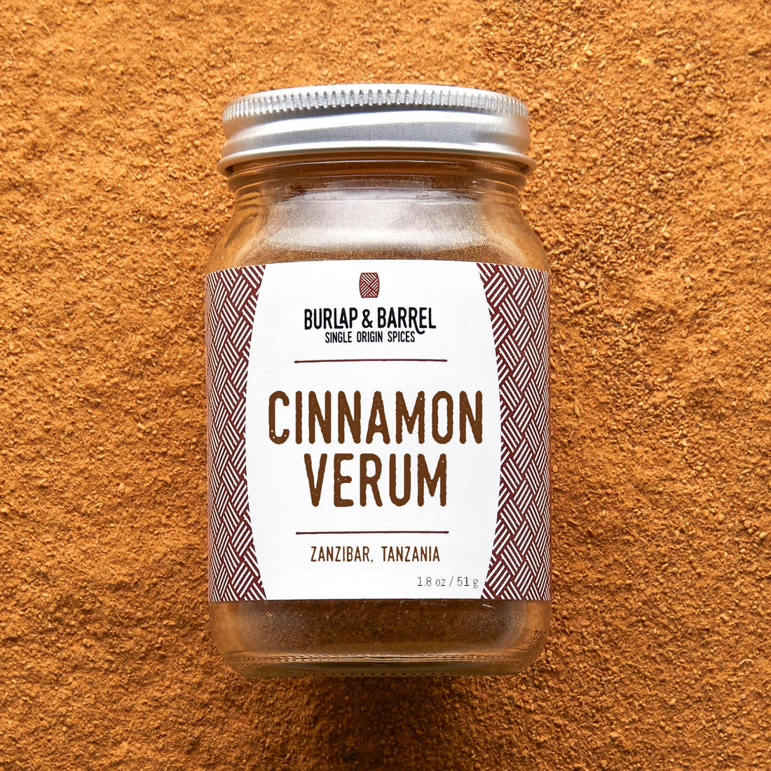 A glass jar of Burlap &amp; Barrel Cinnamon Verum spices, featuring a white label with brown text and patterned side borders, sealed with a silver metal lid and set against a textured orange-brown background.