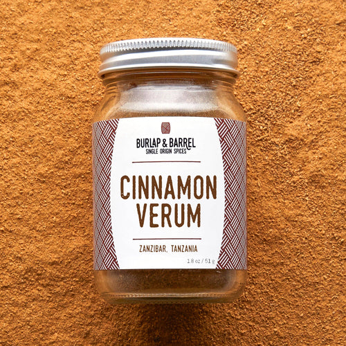 A glass jar of Burlap & Barrel Cinnamon Verum spices, featuring a white label with brown text and patterned side borders, sealed with a silver metal lid and set against a textured orange-brown background.