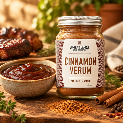 A jar of Burlap &amp; Barrel Cinnamon Verum spice blend stands on a rustic wooden table, surrounded by cinnamon sticks, a bowl of barbecue sauce, and scattered ground cinnamon, with blurred food and greenery in the background.