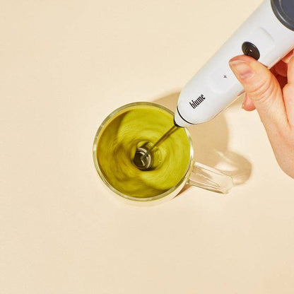 Hand using a white handheld milk frother to create foam in a glass of green liquid on a beige background