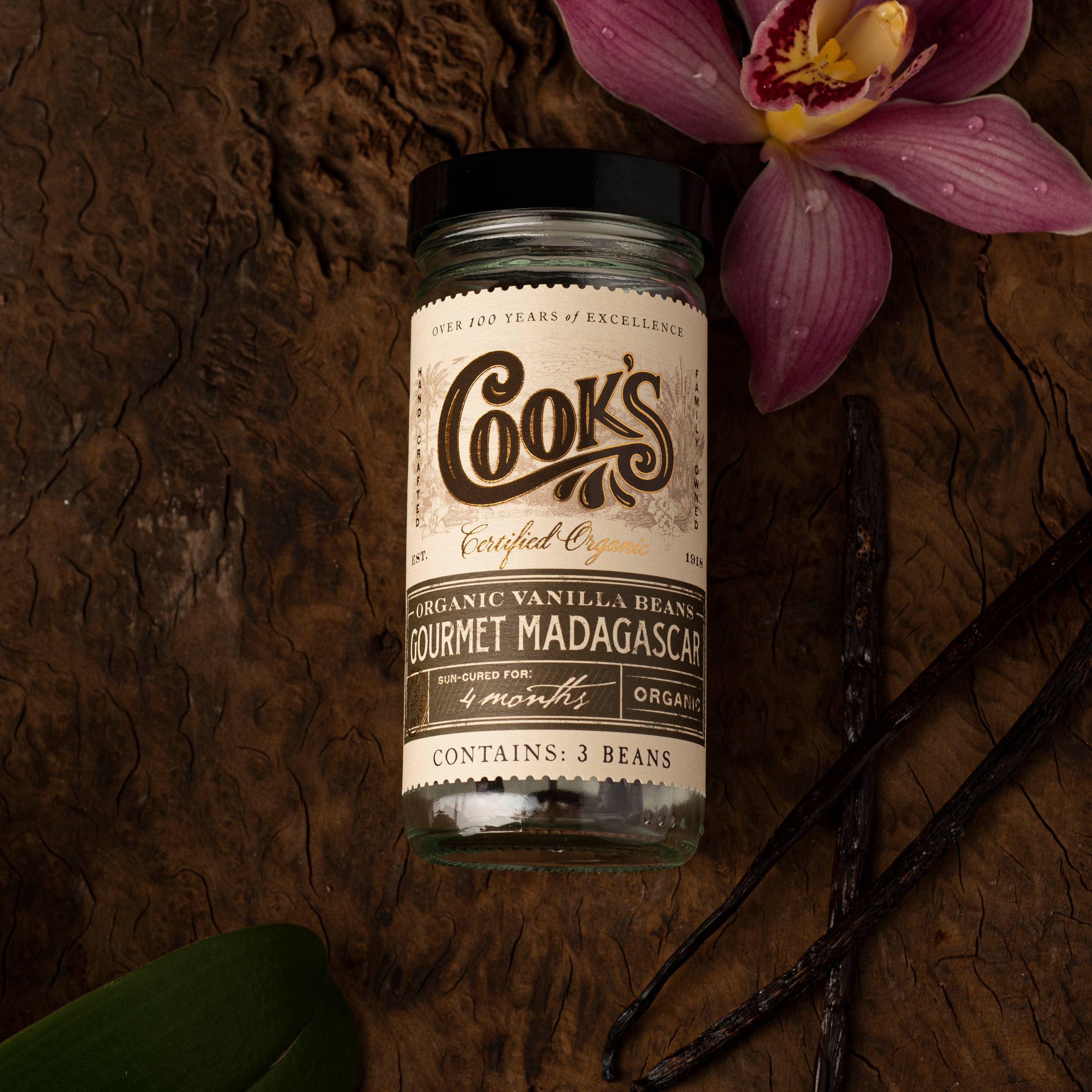 Jar of Cooks Madagascar vanilla beans on a wooden surface with vanilla pods and an orchid.