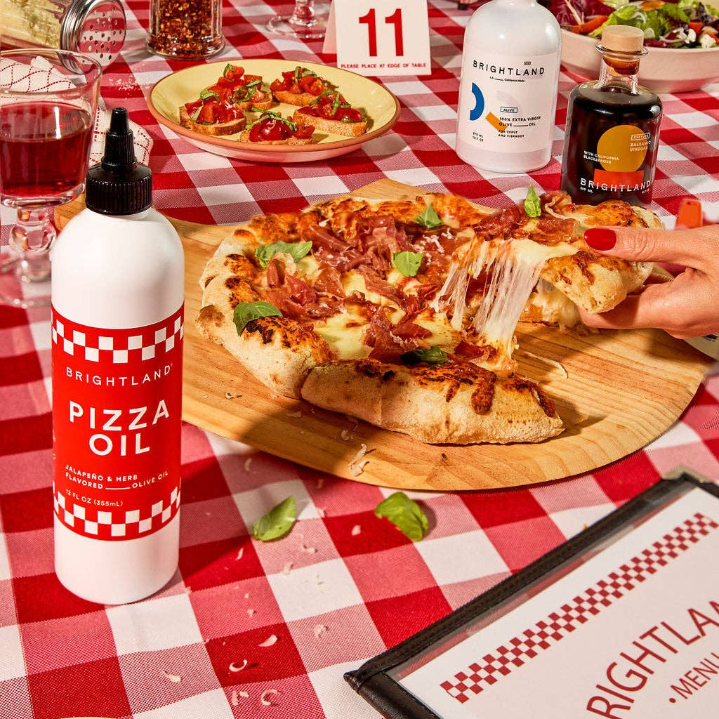 Pizza on a wooden board with Brightland Pizza Oil bottle on a red and white checkered tablecloth.