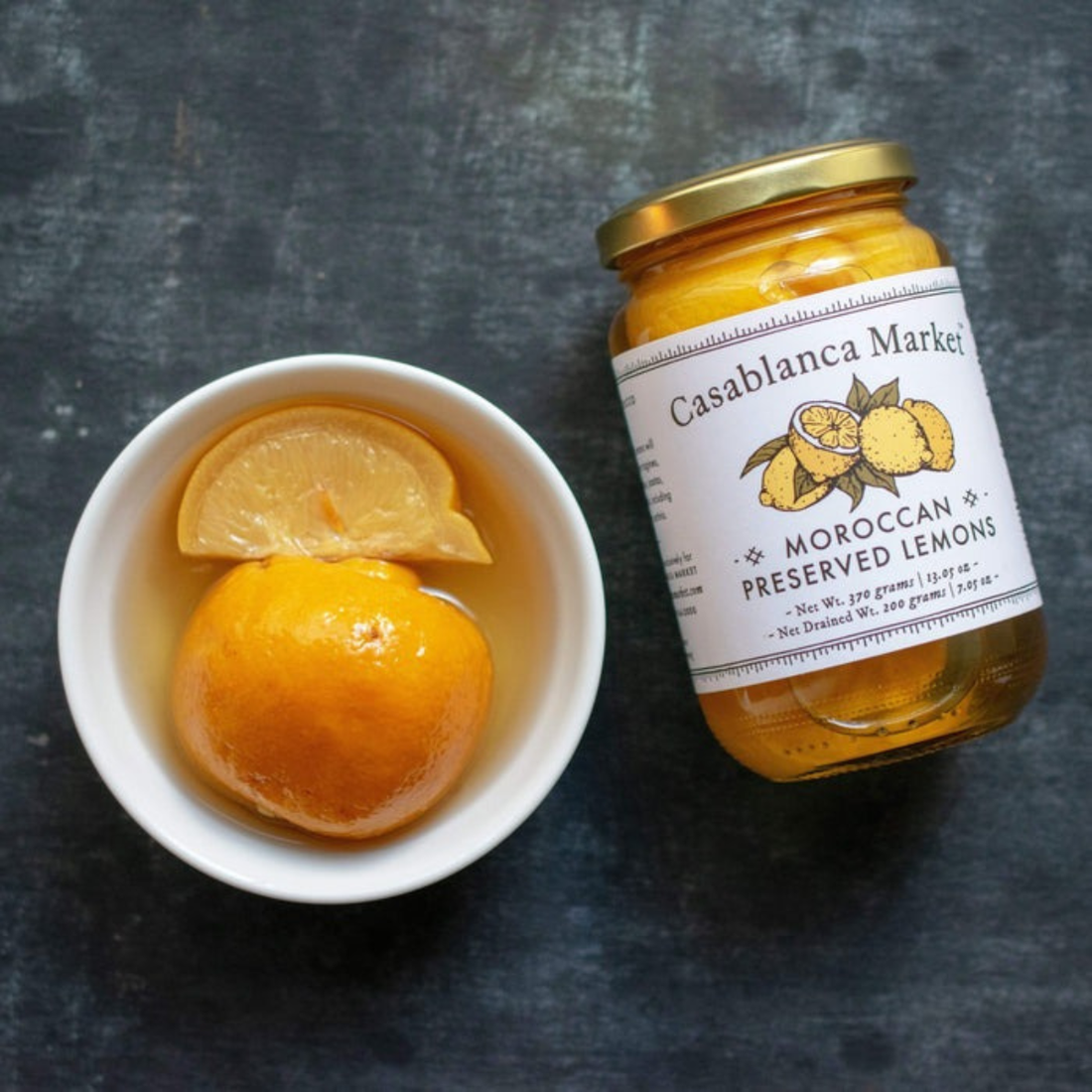 Jar of Casablanca Market Moroccan Preserved Lemons next to a bowl with preserved lemons on a dark surface