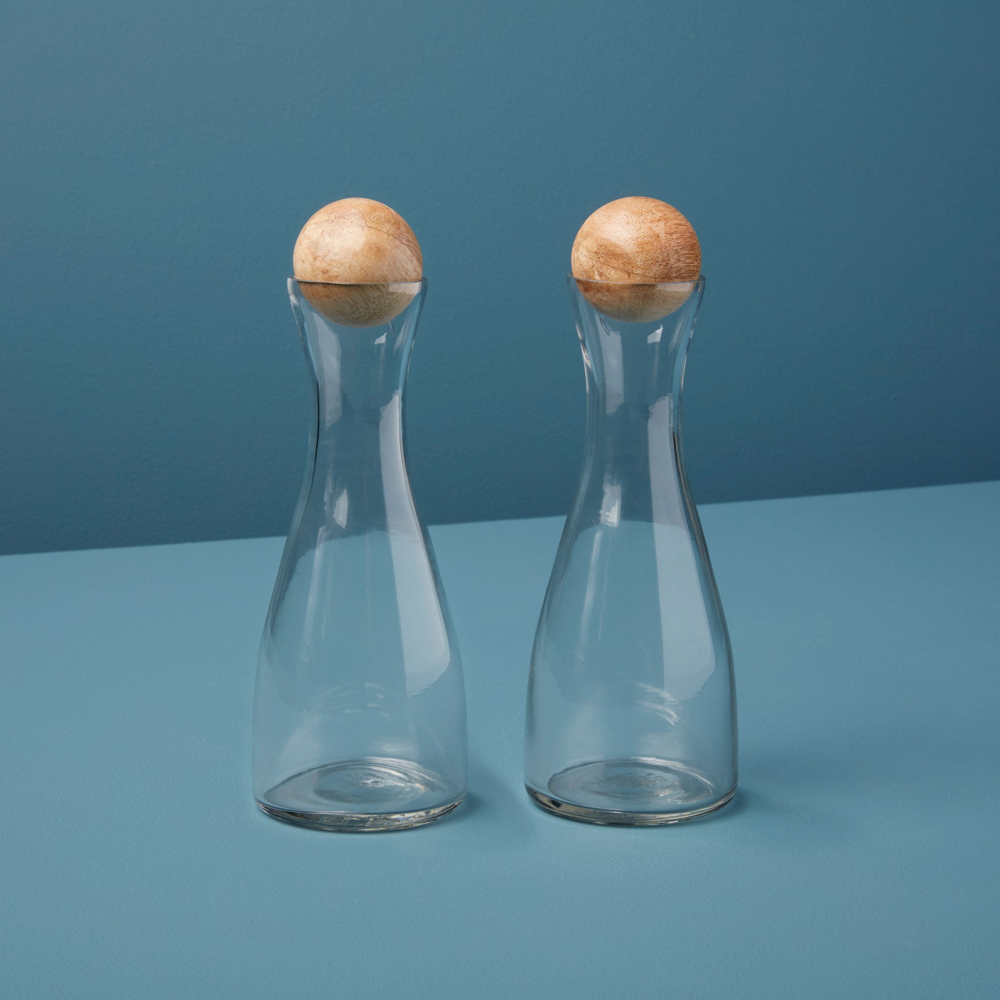 Two clear glass cruets with wooden tops on a blue background
