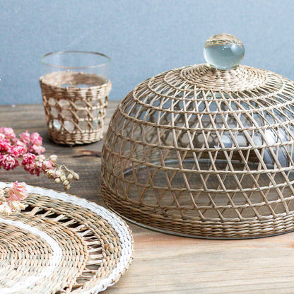 Glass and seagrass cloche on a wooden table with a glass container and flowers in the background.