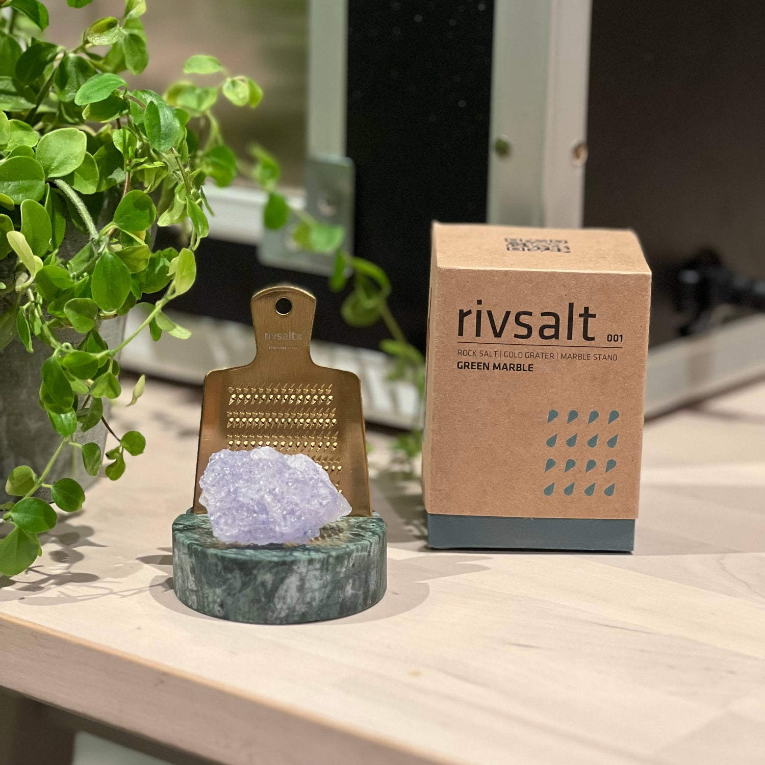 Decorative salt grinder with crystal on a marble stand next to a Rivsalt box on a wooden surface.