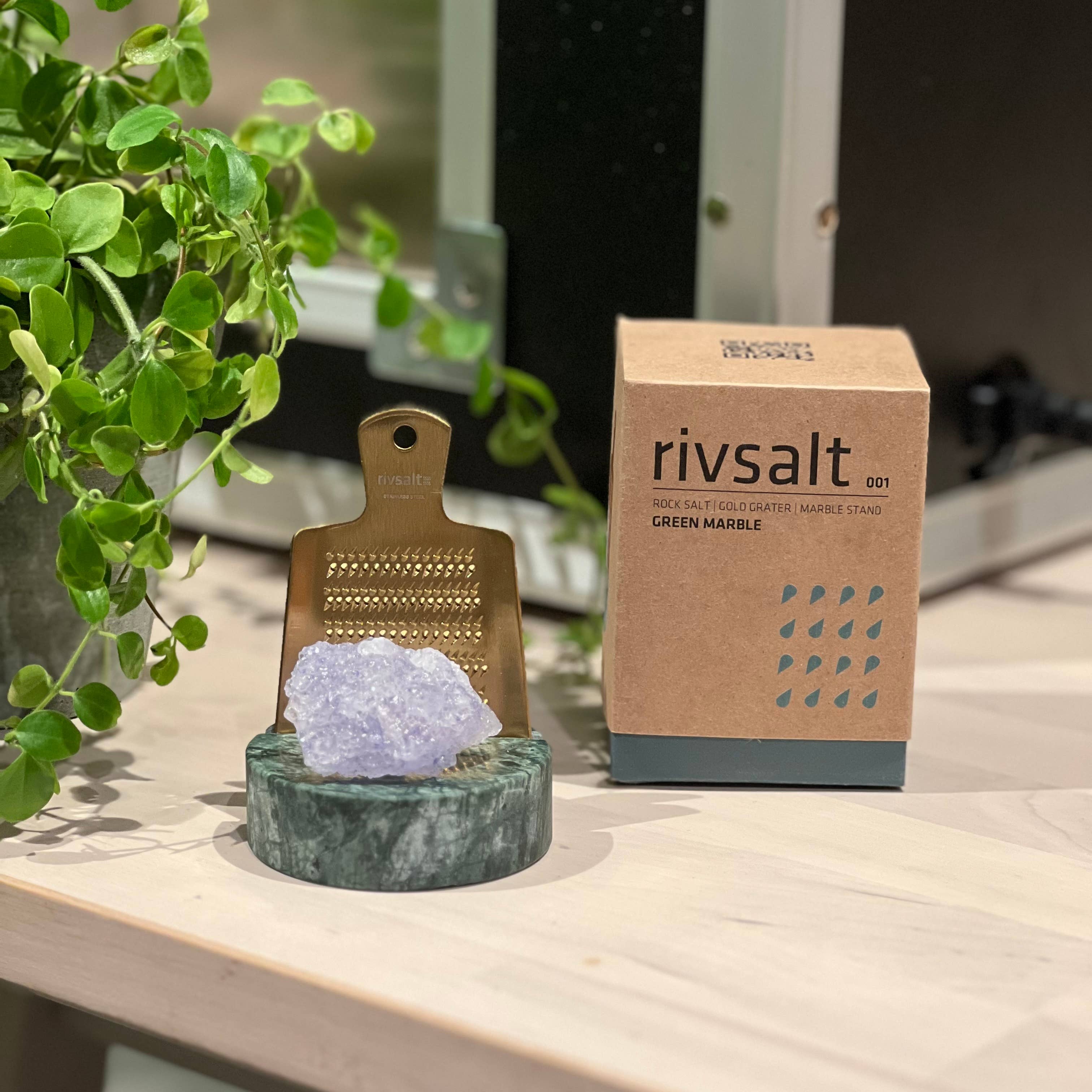 Decorative salt grinder with crystal on a marble stand next to a Rivsalt box on a wooden surface.