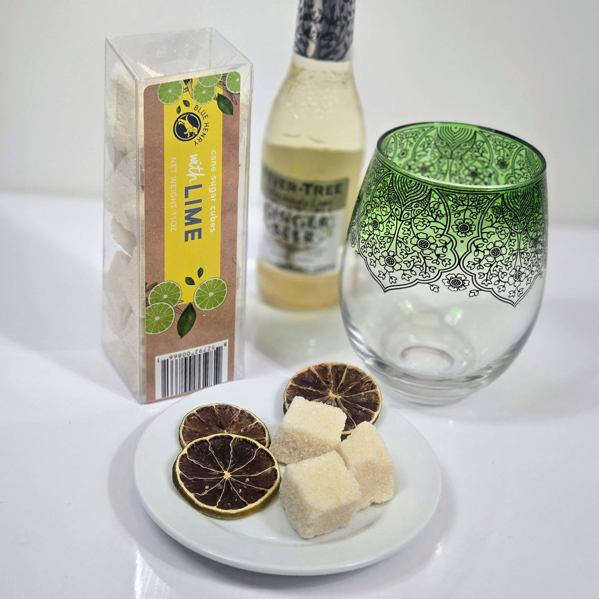 A set of lime-flavored sugar cubes shown with their packaging box, a bottle of ginger beer, and a green patterned glass on a white surface. A small white plate holds dried lime slices and several sugar cubes.