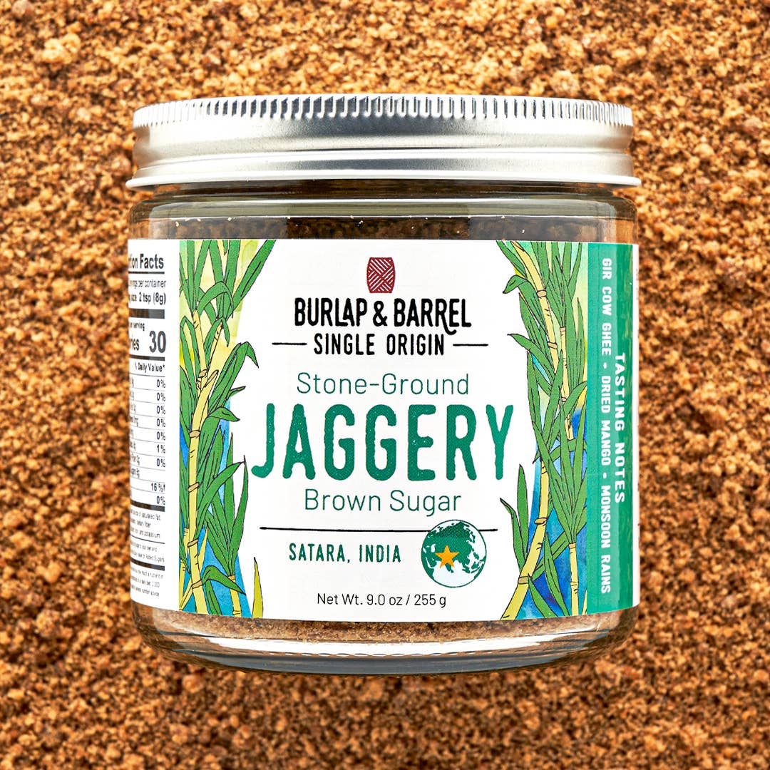 A jar of Burlap &amp; Barrel Stone-Ground Jaggery Brown Sugar, featuring a white label with green illustrations of jaggery leaves, set against a textured brown background of loose jaggery granules.