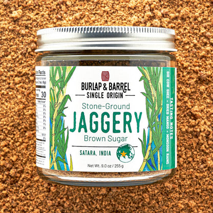 A jar of Burlap &amp; Barrel Stone-Ground Jaggery Brown Sugar, featuring a white label with green illustrations of jaggery leaves, set against a textured brown background of loose jaggery granules.