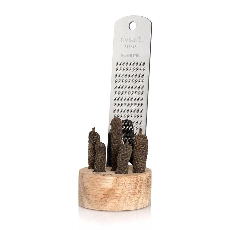 A stainless steel grater set with dark brown textured Java peppers, displayed on a light wood stand against a white background. 