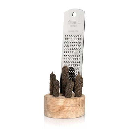 A stainless steel grater set with dark brown textured Java peppers, displayed on a light wood stand against a white background. 