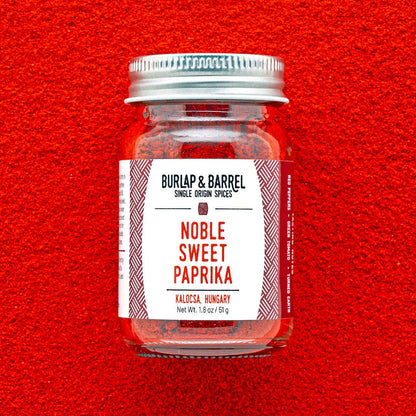 A glass jar of Burlap &amp; Barrel Noble Sweet Paprika centered against a bright red textured background. The jar has a silver metal lid and a white label with red text and geometric patterns.
