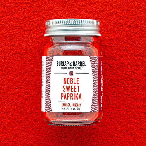 A glass jar of Burlap & Barrel Noble Sweet Paprika centered against a bright red textured background. The jar has a silver metal lid and a white label with red text and geometric patterns.