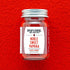 A glass jar of Burlap & Barrel Noble Sweet Paprika centered against a bright red textured background. The jar has a silver metal lid and a white label with red text and geometric patterns.