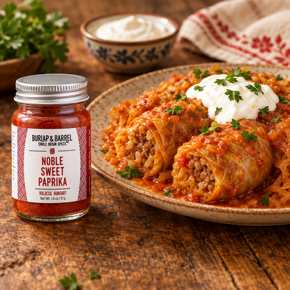 A jar of Burlap &amp; Barrel Noble Sweet Paprika spice placed beside a plated dish of stuffed cabbage on a rustic wooden table, with a bowl of sour cream and a folded cloth in the background.