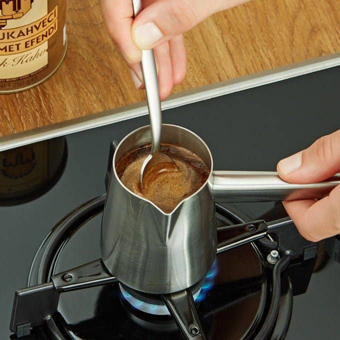 Person using a spoon to stir coffee in a metal pot on a stove with a coffee can labeled &