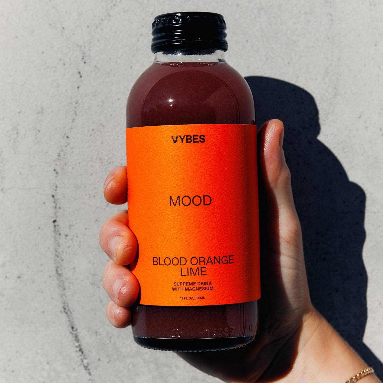 Hand holding a bottle of VYBES Blood Orange Lime juice against a gray background