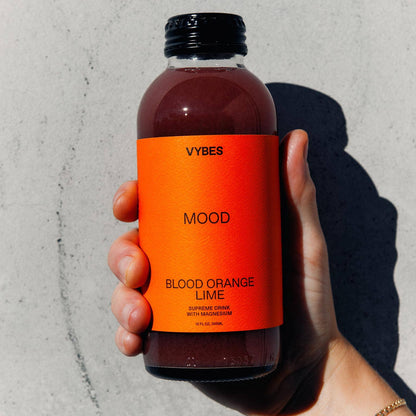 Hand holding a bottle of VYBES Blood Orange Lime juice against a gray background