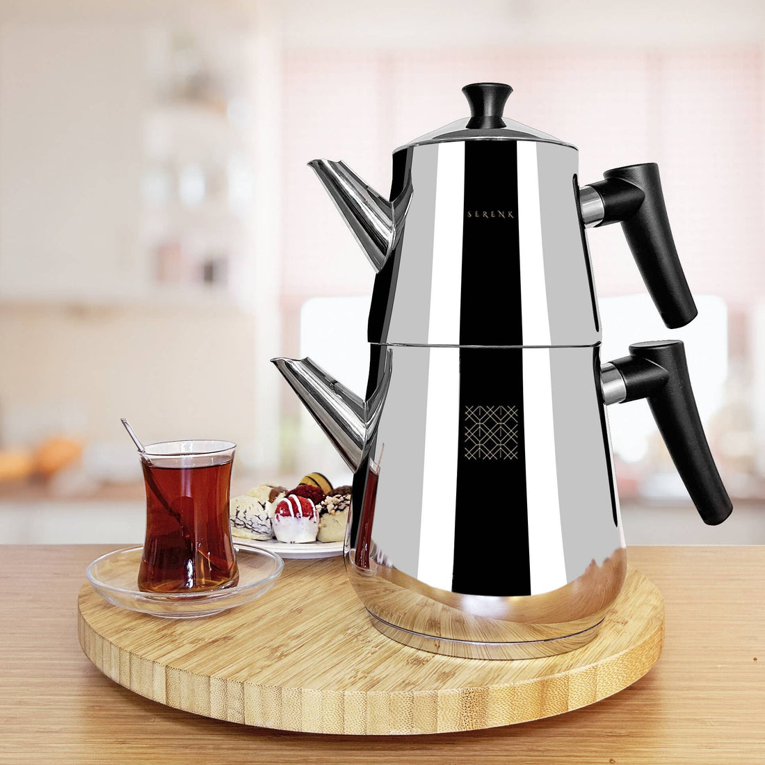 Stainless steel espresso maker on a wooden tray with a glass of tea and snacks in the background.
