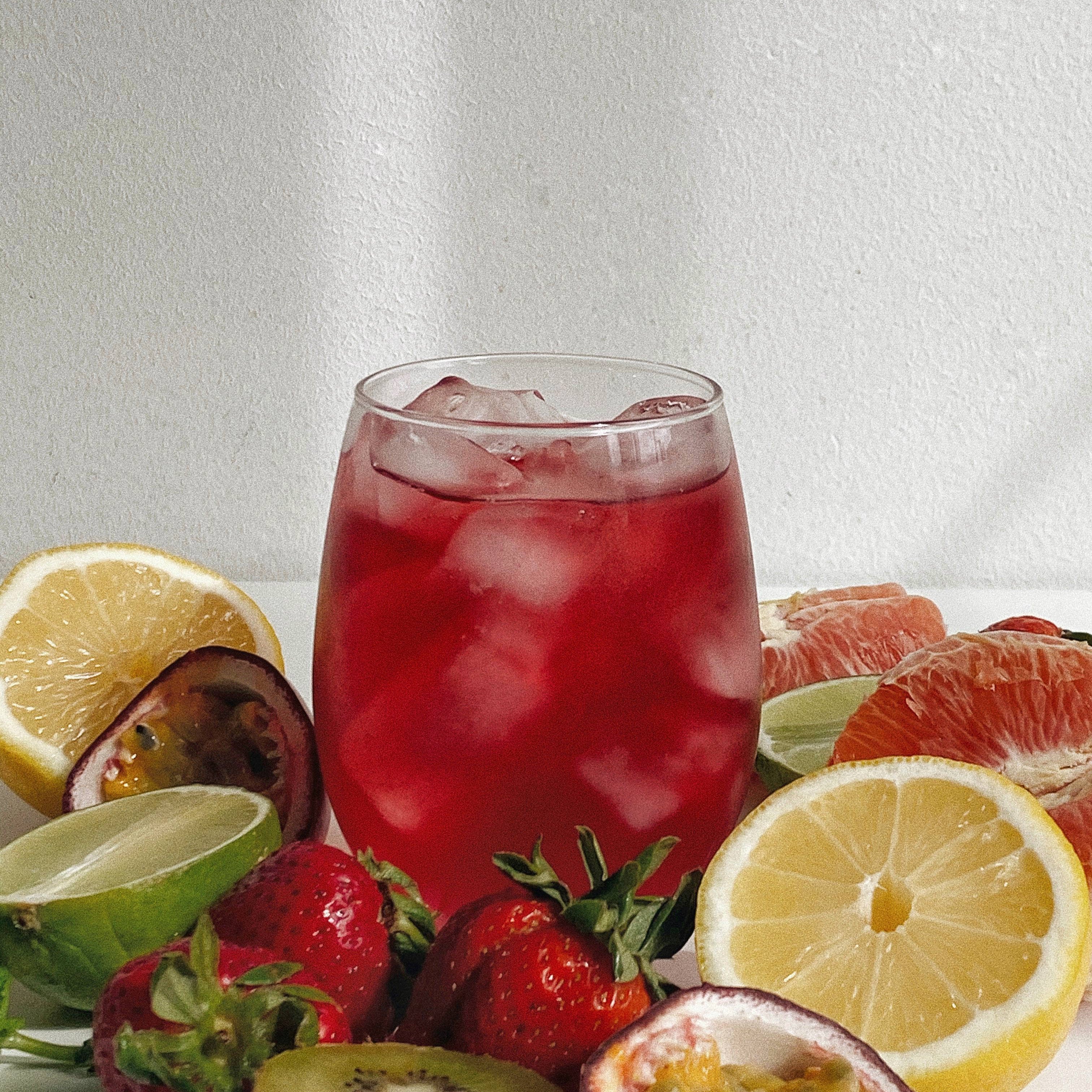 Glass of red juice with ice surrounded by fruits on a white background