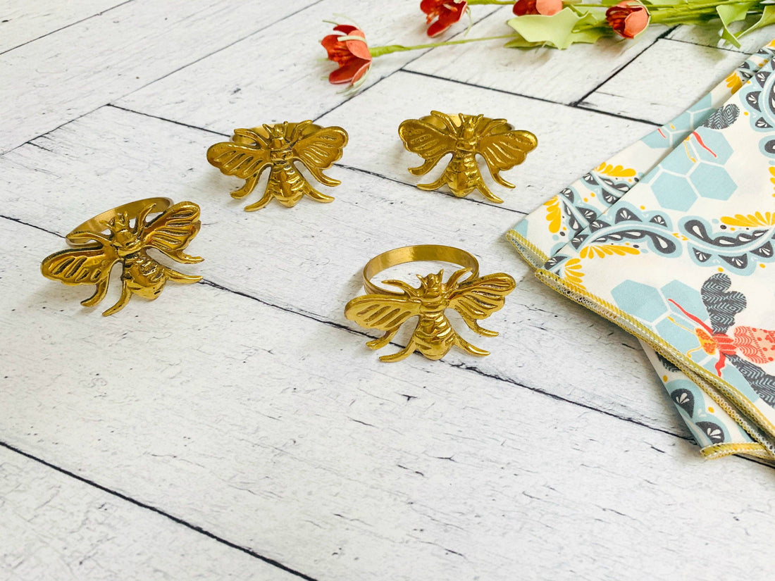 Gold bee-shaped napkin rings on a white wooden surface with honey bee cloth napkins