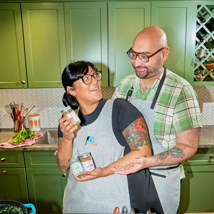 Two people in a kitchen holding jars, with green cabinets and a wine rack in the background.