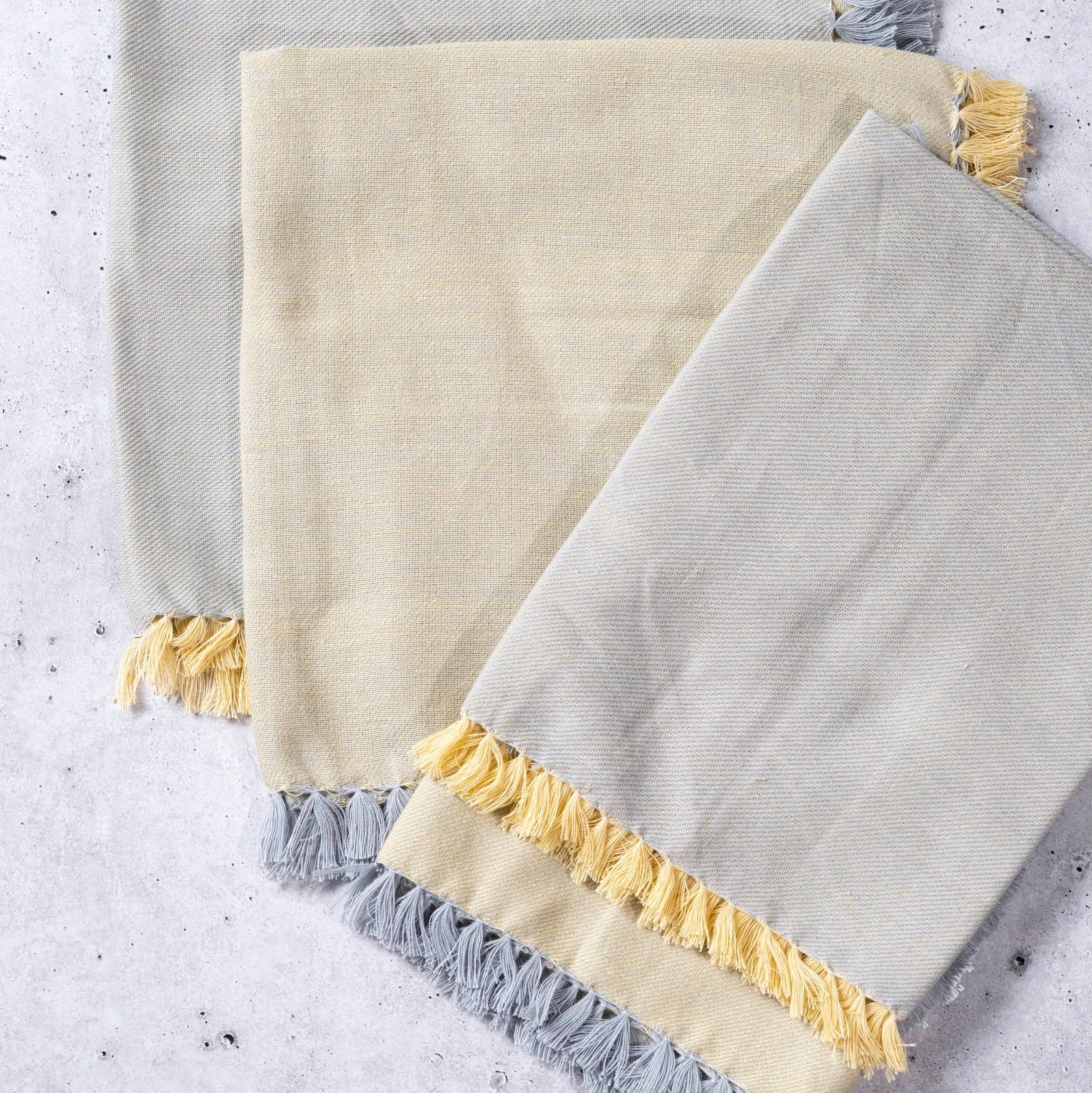 Three textured towels with frayed edges in gray, beige, and yellow on a light gray background.