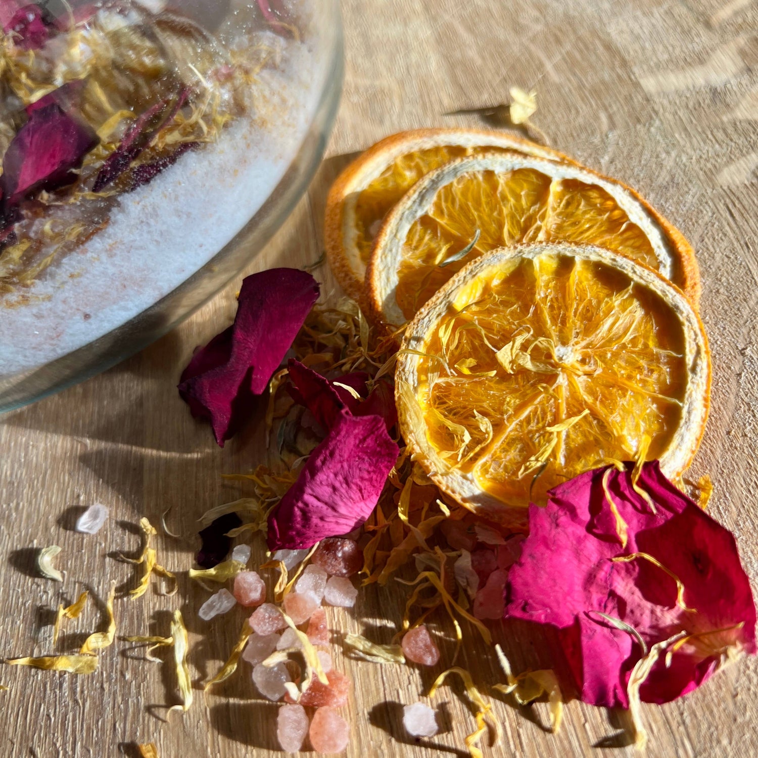 Dried orange slices and pink petals on a wooden surface with a glass container in the background.