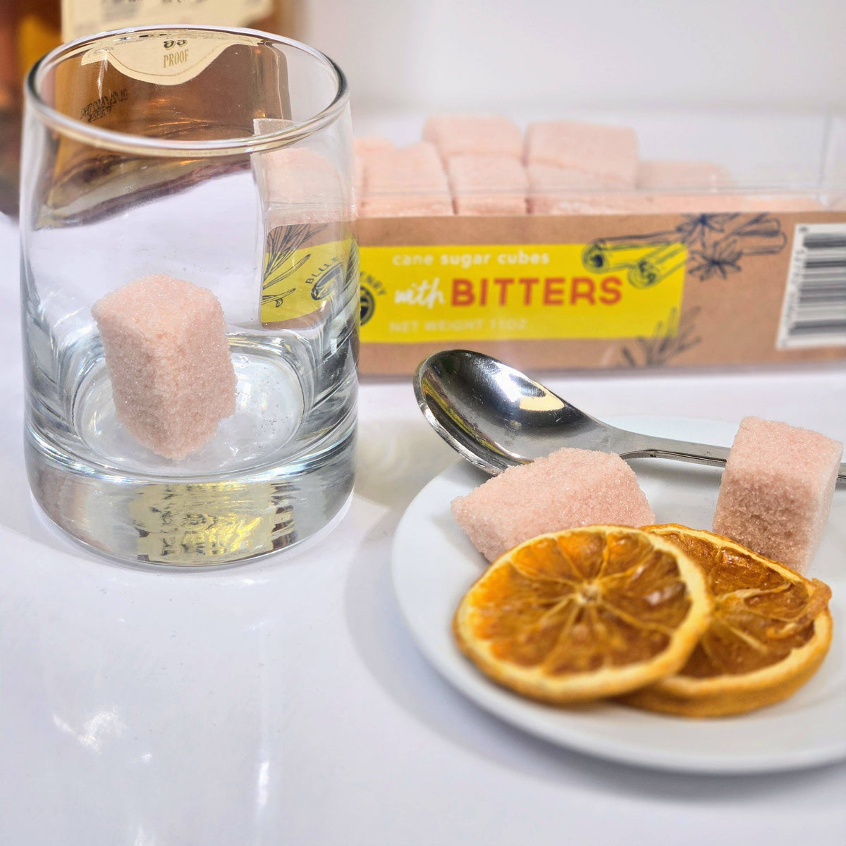 An empty glass with a pink bitters sugar cube inside, alongside a spoon, dried orange slices on a plate, and a box of cane sugar cubes with bitters.