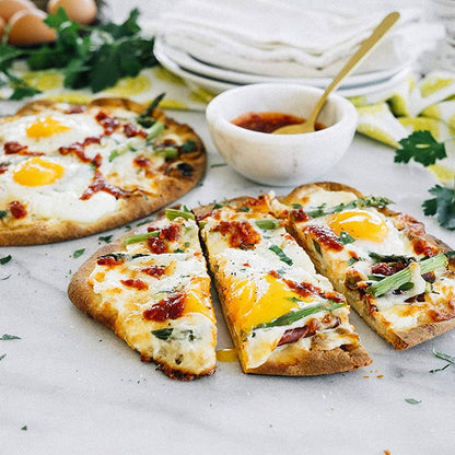 Heart-shaped pizza with eggs and bacon on a marble surface with ingredients in the background.