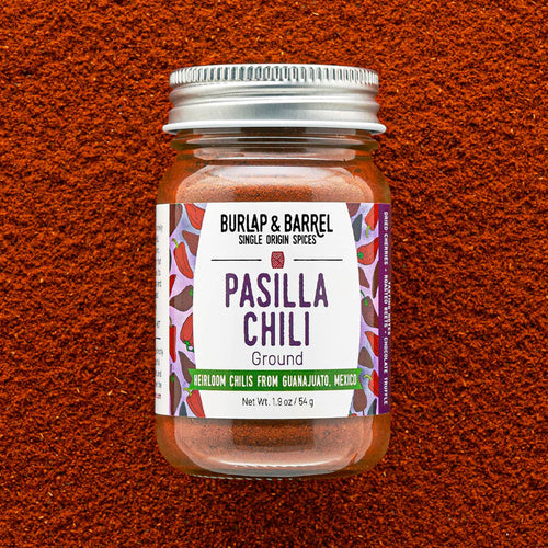 A glass jar of Burlap & Barrel Pasilla Chili Ground, filled with reddish-brown chili powder and topped with a silver metal lid, set against a textured background of the same chili powder.