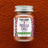 A glass jar of Burlap & Barrel Pasilla Chili Ground, filled with reddish-brown chili powder and topped with a silver metal lid, set against a textured background of the same chili powder.