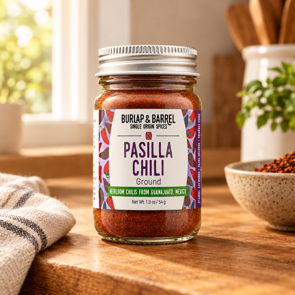 A jar of Burlap &amp; Barrel Pasilla Chili Ground spices sits on a wooden table in a warm kitchen setting, with a striped cloth, a bowl of chili powder, and wooden utensils blurred in the background.