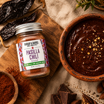 A glass jar of Buriap &amp; Barrel Pasilla Chili Ground spice with a silver lid and colorful label, styled on a wooden cutting board next to a bowl of mole sauce and dried chilies.