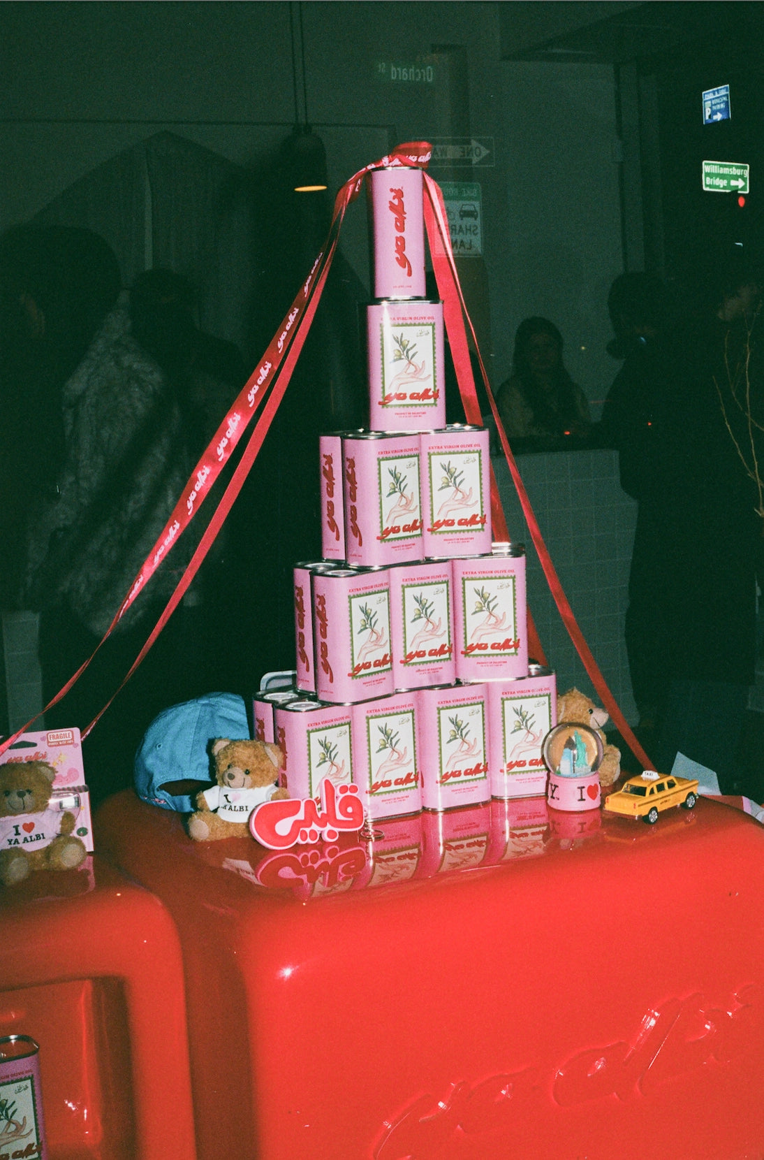 Pyramid of Ya Albi olive oil cans with teddy bears on a red surface, blurred background