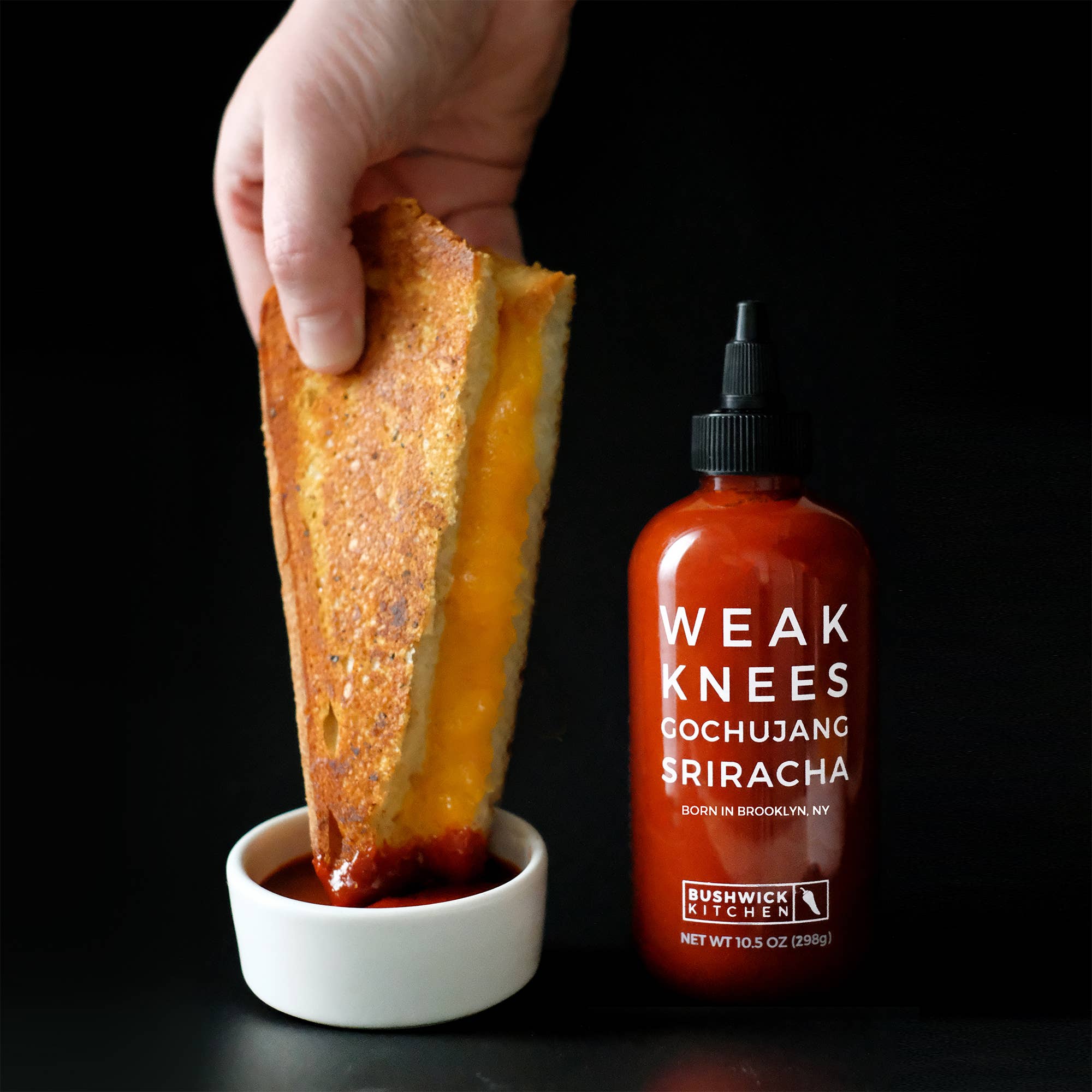Hand dipping a grilled cheese sandwich into a bowl of sauce next to a bottle of &
