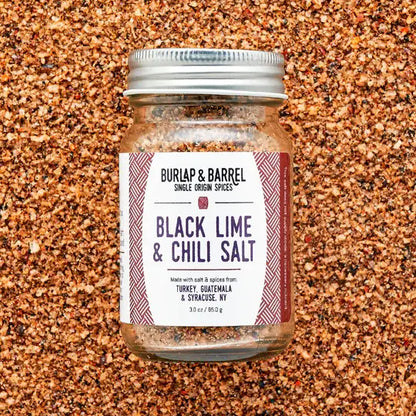 A glass jar of Burlap &amp; Barrel Black Lime &amp; Chili Salt, filled with coarse brown and reddish-brown spices, set against a background of the same spice blend. The jar has a silver metal lid and a white label with dark purple text and red patterned borders.