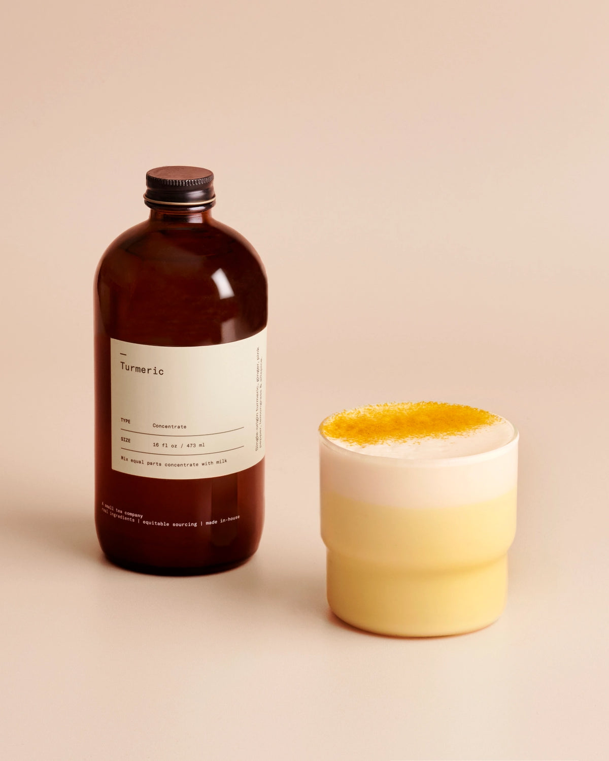 An amber glass bottle Turmeric Concentrate stands next to a frosted glass containing a golden milk latte topped with a golden sprinkle, set against a solid beige background.