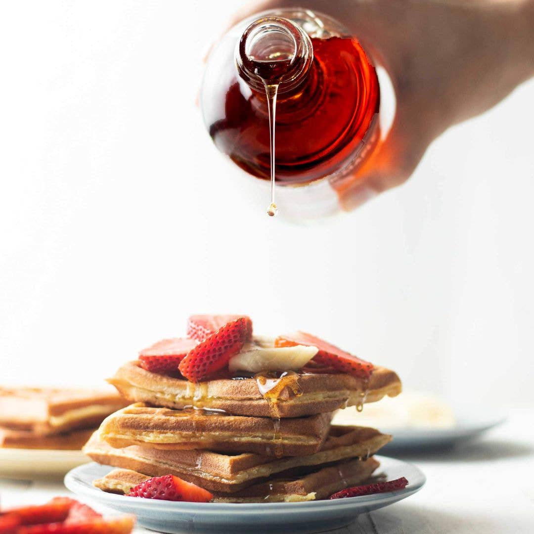 Stack of waffles with strawberries being drizzled with syrup on a white plate.