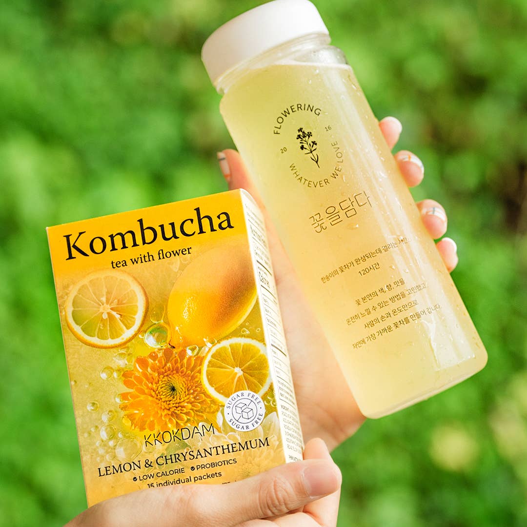 Hand holding a bottle and box of Kombucha tea with lemon and chrysanthemum against a green blurred background.