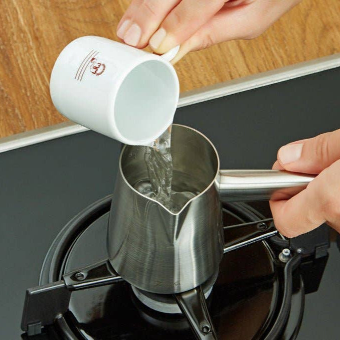 Person pouring water from a white cup into a metal pot on a stove.