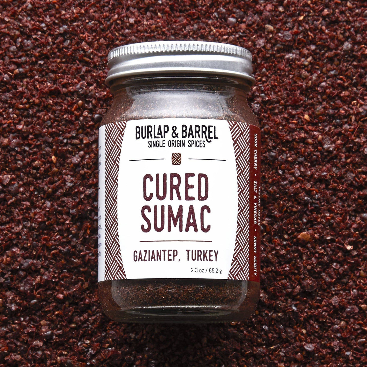 A glass jar of Burlap &amp; Barrel Cured Sumac (Gaziantep, Turkey) with a white label and silver lid, set against a textured background of dark brown cured sumac spice.