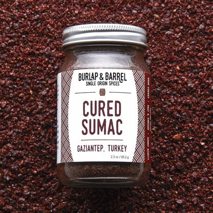 A glass jar of Burlap &amp; Barrel Cured Sumac (Gaziantep, Turkey) with a white label and silver lid, set against a textured background of dark brown cured sumac spice.